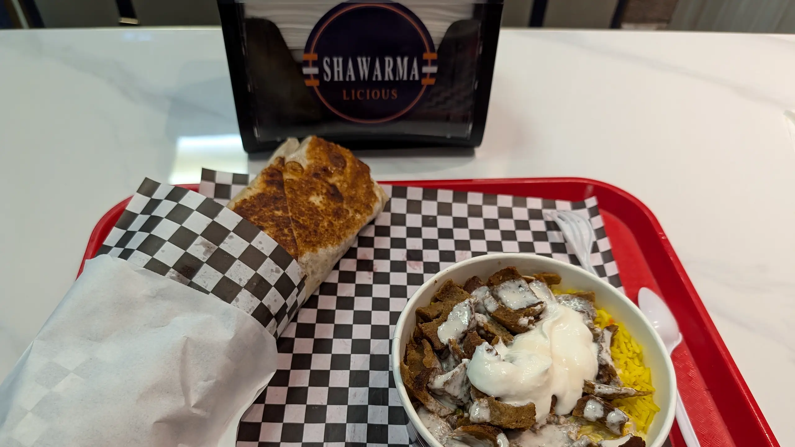 Beef donair over yellow rice with garlic sauce and a toasted wrap on a checkered tray, with a Shawarmalicious napkin holder behind