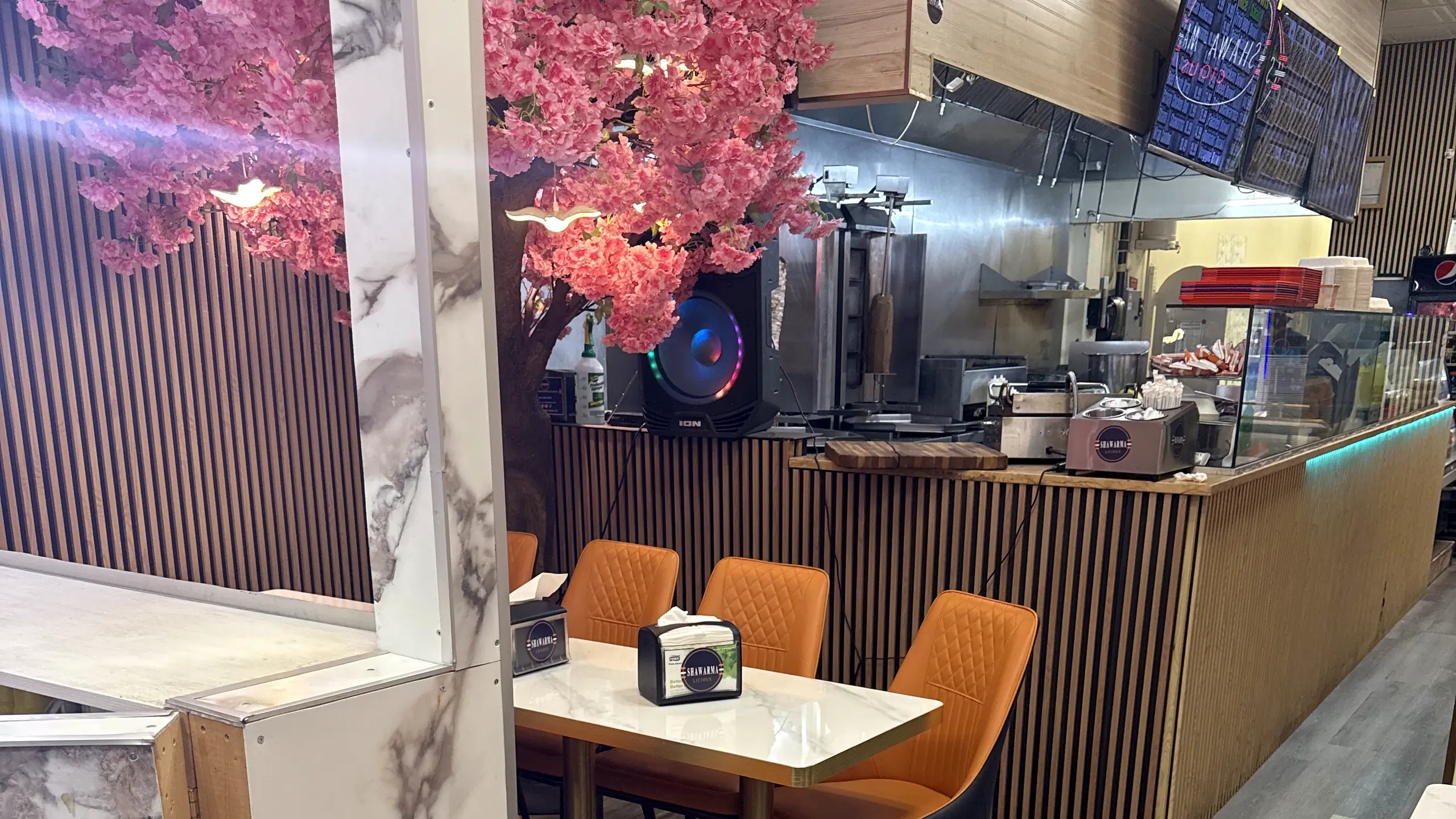 Dining room with orange chairs at marble tables, a pink blossom tree and the shawarma counter