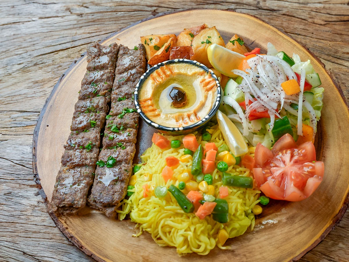 Two beef kabab skewers with yellow rice, hummus, garlic potatoes and salad on a wooden board