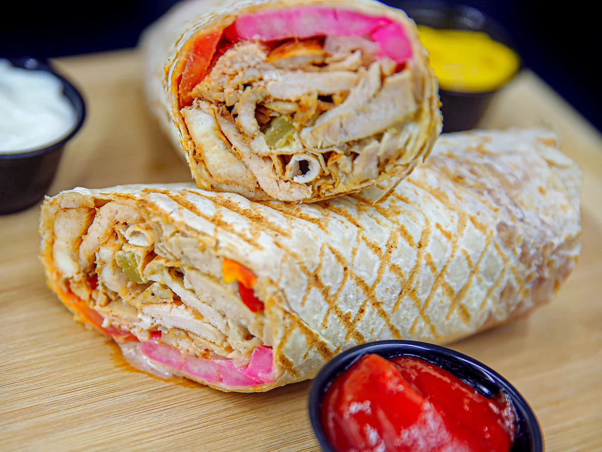 Chicken shawarma wrap cut in half, filled with chicken, pickles and pink turnip, with garlic sauce and hot sauce cups